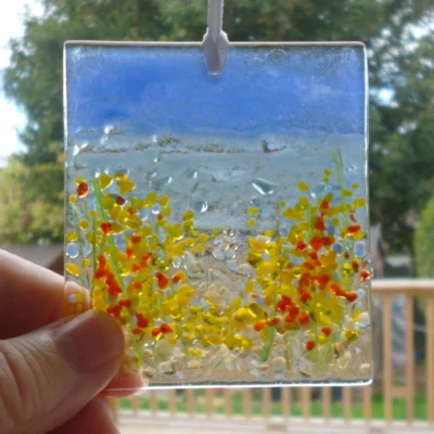 Fused glass coastal suncatcher hanging in a window being held by the bottom left corner. Natural light shining through showing detail.