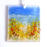 Handmade rectangle fused glass coastal suncatcher laying on a white background. Red, yellow and blue flowers on a beach. Hanging from a white ribbon.
