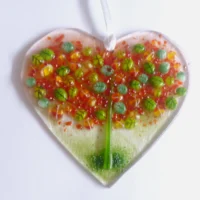 A very tactile fused glass heart suncatcher with autumn tree in colours of green, red and orange, hanging from a white ribbon.