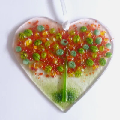 A very tactile fused glass heart suncatcher with autumn tree in colours of green, red and orange, hanging from a white ribbon.