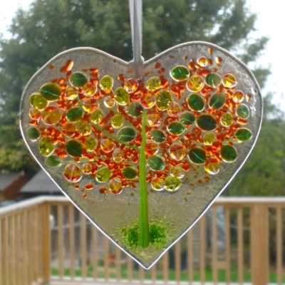Hand cut fused glass heart suncatcher with an autumn tree in green and red, hanging from a white ribbon.