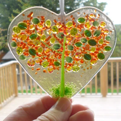Hand cut fused glass heart suncatcher hanging in a window being held at the bottom to show size.