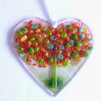 Fused glass heart suncatcher with autumn tree on top, with green and red leaves with green trunk. Hole at the top with white ribbon threaded through to hang from.