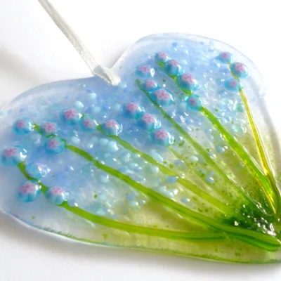 Fused glass heart suncatcher laying at an angle showing detail of glass flowers and stems, white ribbon threaded through the top to hang from.