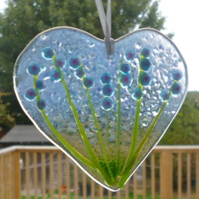 Hand cut fused glass heart suncatcher with blue flowers hanging in a window from a white ribbon.