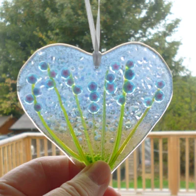 Fused glass heart suncatcher with blue flowers and green stems being held at the bottom. Hanging from a white ribbon