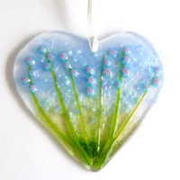 Fused glass heart suncatcher with blue and pink flowers and green stems, hanging from a white ribbon.