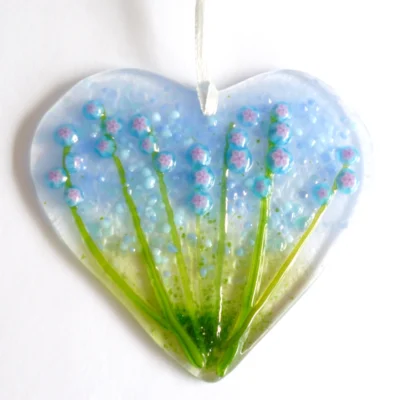 Fused glass heart suncatcher with blue and pink flowers and green stems, hanging from a white ribbon.