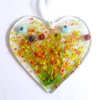Quirky fused glass heart suncatcher with square flowers in red, blue and yellow, hanging from a white ribbon. Colourful pieces of yellow, red and orange fused on top,