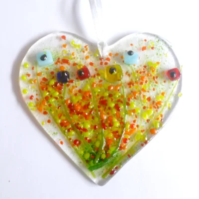 Quirky fused glass heart suncatcher with square flowers in red, blue and yellow, hanging from a white ribbon. Colourful pieces of yellow, red and orange fused on top,