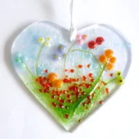 Fused glass heart suncatcher with colourful flowers in red, yellow, orange, blue, lilac and red, laying on a white background.
