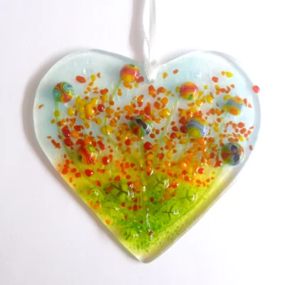 Fused glass heart suncatcher with colourful flowers on top, laying on a white background. Hangs from a white ribbon.