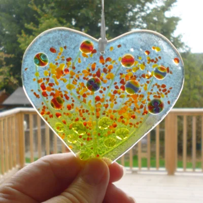 Fused glass heart suncatcher colourful quirky flowers in red, blue, yellow and green. Suncatcher being held at the bottom to show size.