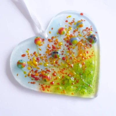 Fused glass heart suncatcher laying on a white ribbon. Colourful quirky flowers in red, blue, and yellow fused on top.