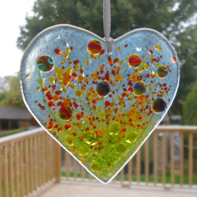 Fused glass heart suncatcher hanging in a window from a white ribbon. Quirky colourful flowers fused onto a clear piece of glass.