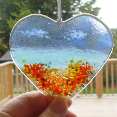 Coastal fused glass suncatcher with a blue background and orange & yellow frit at the bottom to represent flowers. Suncatcher being held to show size.