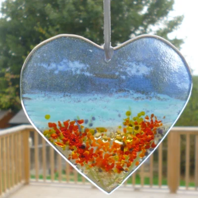 Fused glass heart suncatcher with coastal and floral scene hanging in a window from a white ribbon.