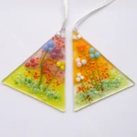 Set of two handmade glass flower suncatchers in a triangle shape with beads of colourful glass to create flowers in red, blue, turquoise, plum, orange, pink, yellow and white. Hole drill at the top point and has a white ribbon threaded through. Green and red creates more decoration. Very tactile and colourful