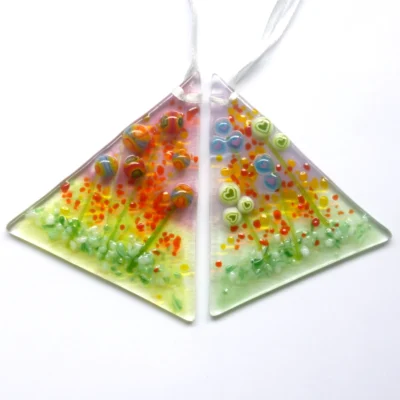 Two handmade fused glass suncatchers with quirky flowers in blue and green heart flowers and red and orange bead flowers. Each suncatcher is hanging from a white ribbon.