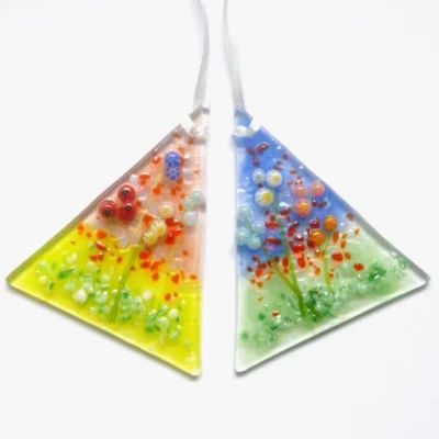 Two small fused glass flower suncatchers hanging from white ribbons. One suncatcher has red, blue, yellow glass beads for flowers with red and green background. Second suncatcher has blue and green background, with cream, blue, and pink glass beads for quirky flowers.