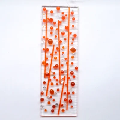 A bubbly orange fused glass suncatcher with orange and dichroic glass beads and stringers fused onto an oblong shaped piece of clear glass. Hanging from a clear cord.
