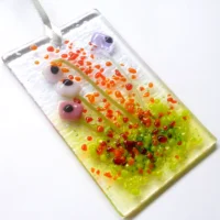 Pink and lilac glass flower suncatcher laying at a slight angle. Pink, lilac and white quirky square glass flowers with green stringers for stems. Red, orange and green frit for decoration.