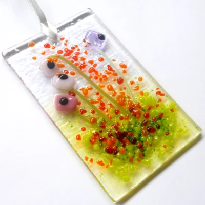 Pink and lilac glass flower suncatcher laying at a slight angle. Pink, lilac and white quirky square glass flowers with green stringers for stems. Red, orange and green frit for decoration.