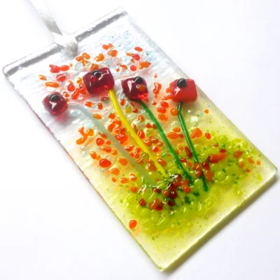 Quirky red glass flower suncatcher laying at a slight angle. Pieces of square red glass has been used to create flowers. Orange and red frit for decoration.