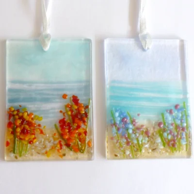 Two small rectangle fused glass coastal suncatchers with blue & red flowers on one, and orange & yellow flowers on the other, laying on a white background.
