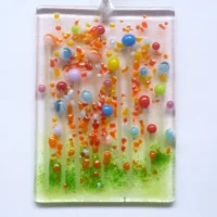 Small rectangle shaped handmade glass flower suncatcher with quirky tactile bead flowers in blue, pink, yellow, green, and pink. Green, orange and red frit decoration. Hangs from a white ribbon.
