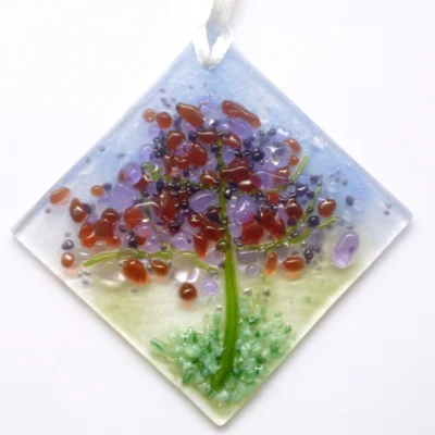 Small spring colours fused glass suncatcher made with the design of a spring tree, and colours of lilac, purple, and plum leaves on a the tree and green stringers for the trunk. The suncatcher hangs from a white ribbon.