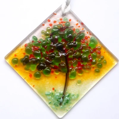 Small summer colours fused glass suncatcher made with the design of a tree in summer time, and colours of red, green and orange and stringers for the trunk and branches. The suncatcher hangs from a white ribbon.
