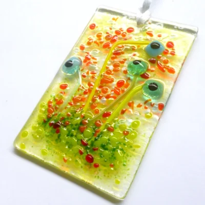 A turquoise glass flower suncatcher laying at a slight angle with a white ribbon attached for hanging. Red, orange and green frit for decoration.